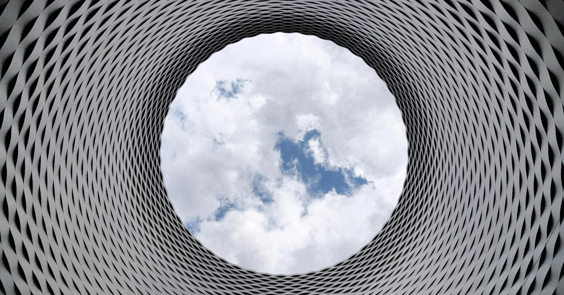 幾何学的な模様を描くビルの内側から、突き抜けるような青空を見上げた構図。複雑な課題を乗り越えた先にある、開かれた未来やビジョンを象徴している。