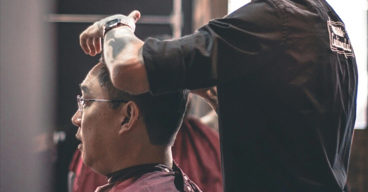 Side profile of a tattooed barber carefully cutting the hair of a male customer with glasses in a Hong Kong barber shop