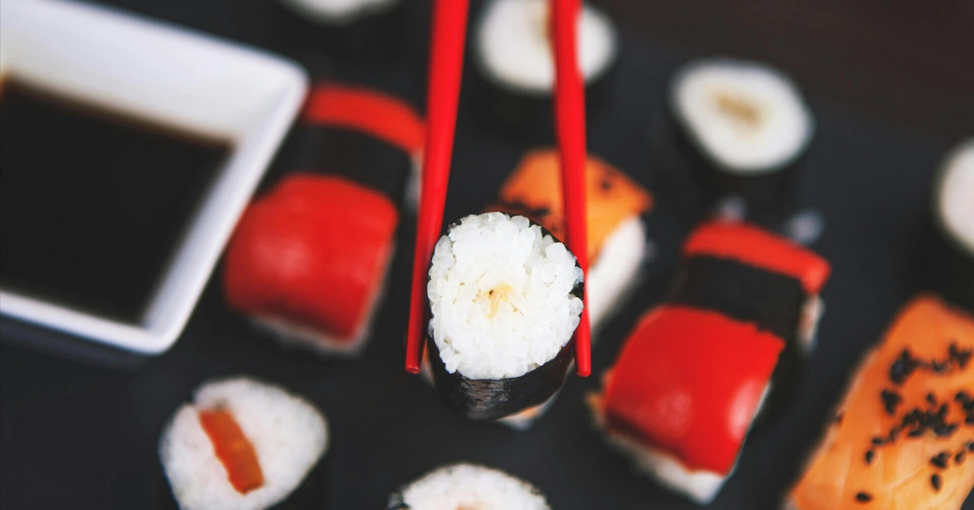 A sushi roll held by red chopsticks with colorful sushi in the background, representing a casual and fun Japanese dining experience