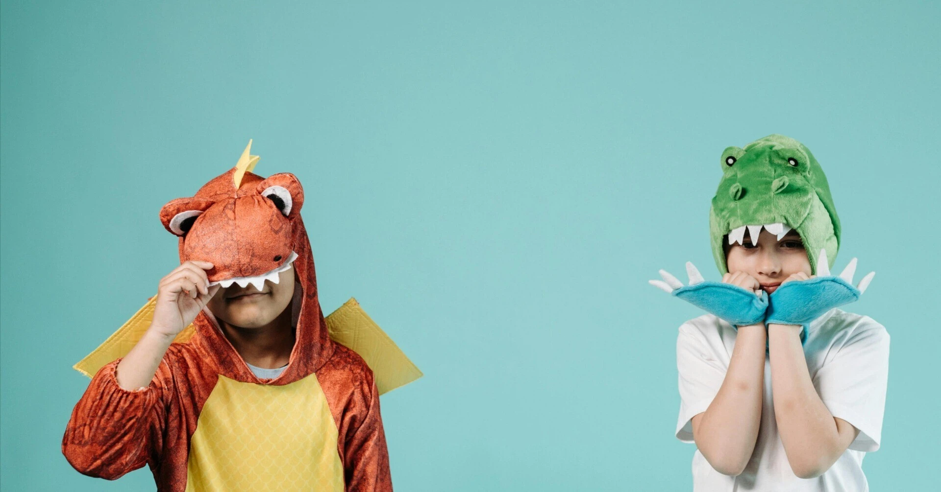 Two children wearing green and orange dinosaur costumes posing playfully against a blue background