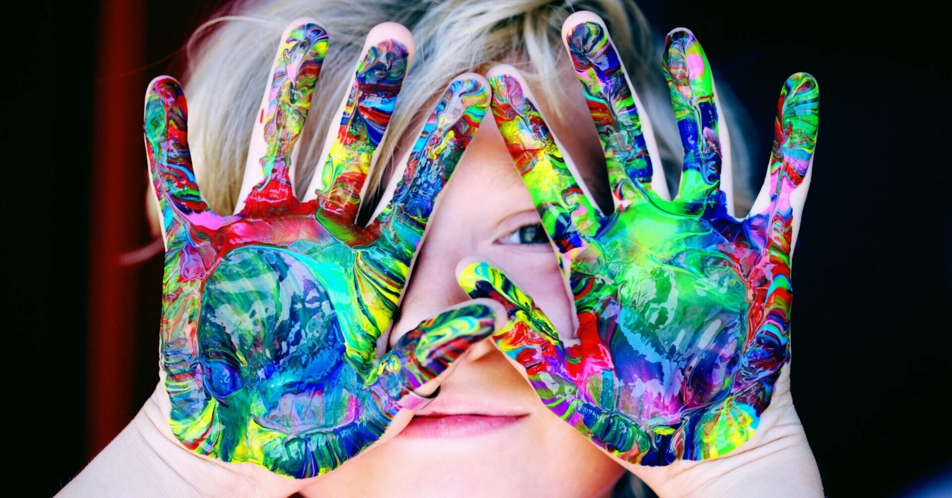 Close-up of a child holding up both hands covered in colorful paint in front of their face, peering through their fingers.