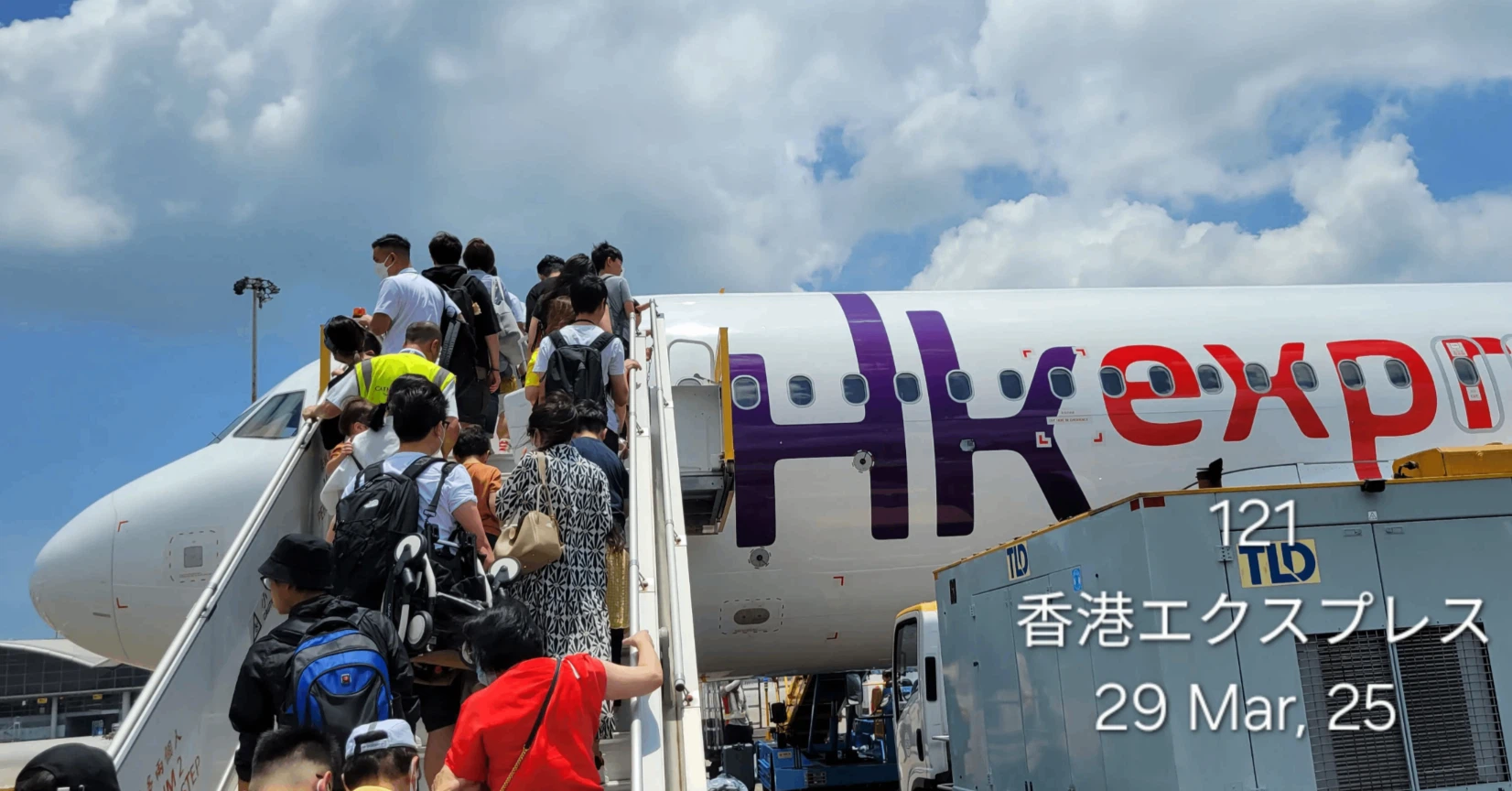 空港の駐機場(タラップ)から、香港エクスプレスの飛行機に搭乗しようとしている乗客の後ろ姿。機体には紫色のロゴが描かれている。