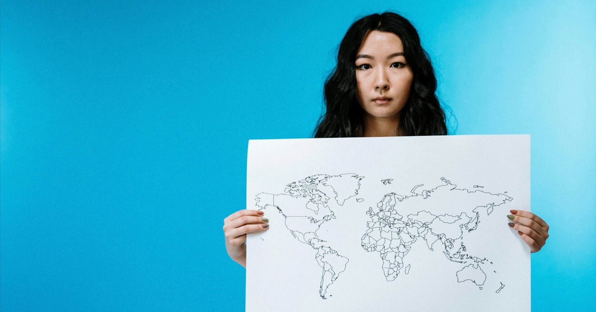 青い背景の前で、世界地図の輪郭が描かれた紙を両手で持つ女性のポートレート。国際市場での戦略的なアプローチを表現。