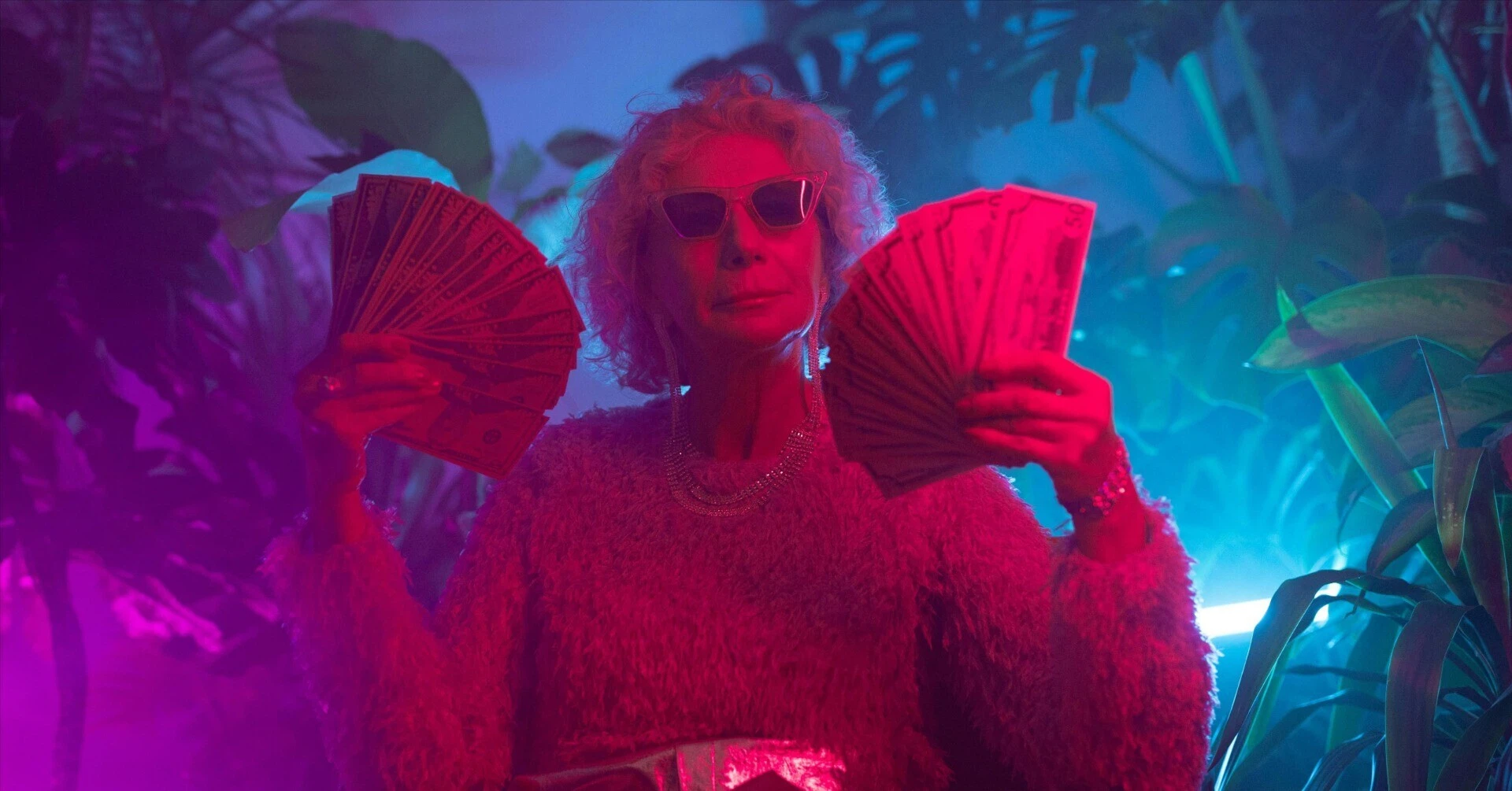 A wealthy older woman wearing sunglasses, holding fans of dollar bills in neon lighting