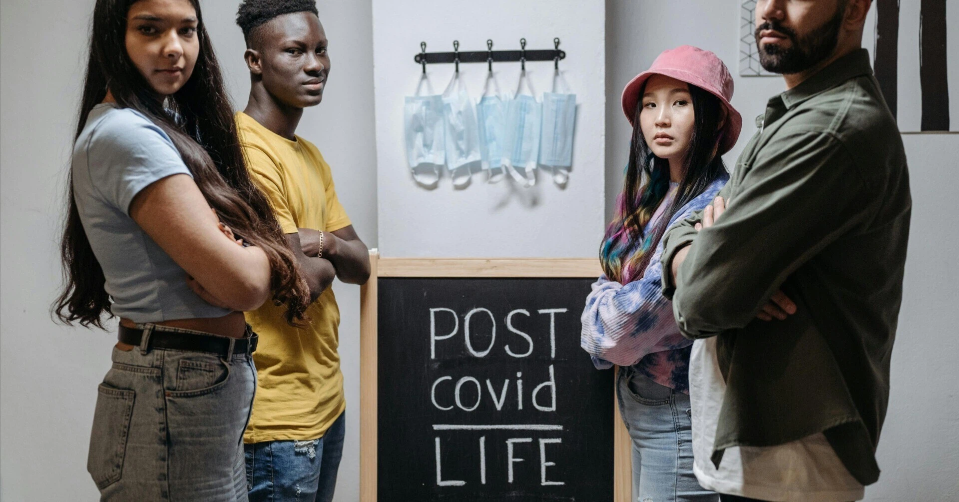 「POST COVID LIFE」と書かれた黒板の周りに立つ若者たちと、壁に吊るされたマスク